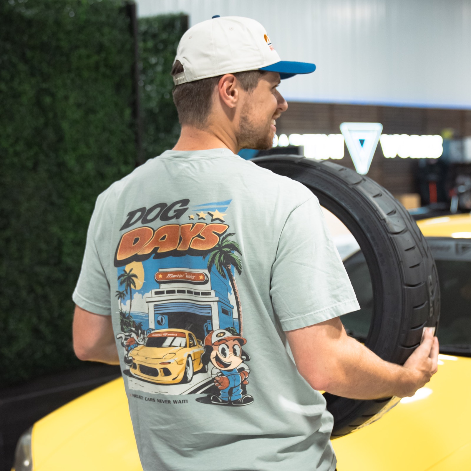 A MartiniWorks team member in a Dog Days tee carrying a Falken tire past a yellow project car